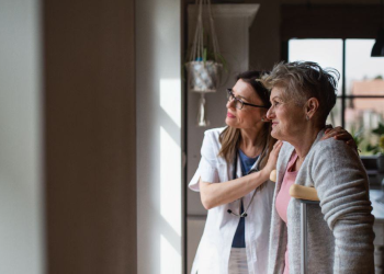 Side view of a healthcare worker or caregiver visiting senior woman indoors at home, helping her to walk.