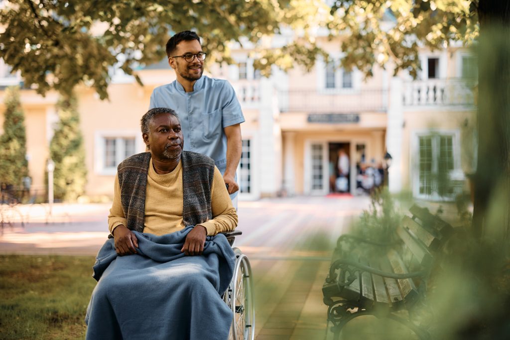 African American senior in wheelchair being pushed by male caregiver in the park of residential care home.