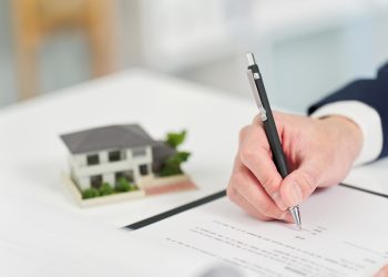 A close-up of a person's hand signing a real estate or building contract with a black pen on a white desk, with a small scale model of a modern house in the background.