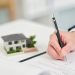 A close-up of a person's hand signing a real estate or building contract with a black pen on a white desk, with a small scale model of a modern house in the background.