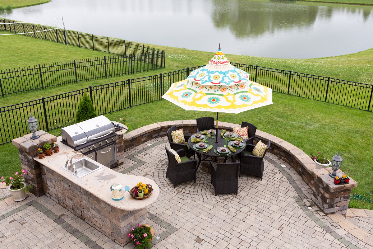 Outdoor living space on a brick patio overlooking a tranquil lake and fenced green lawn with a table under a sunshade or umbrella laid ready for dinner, high angle view