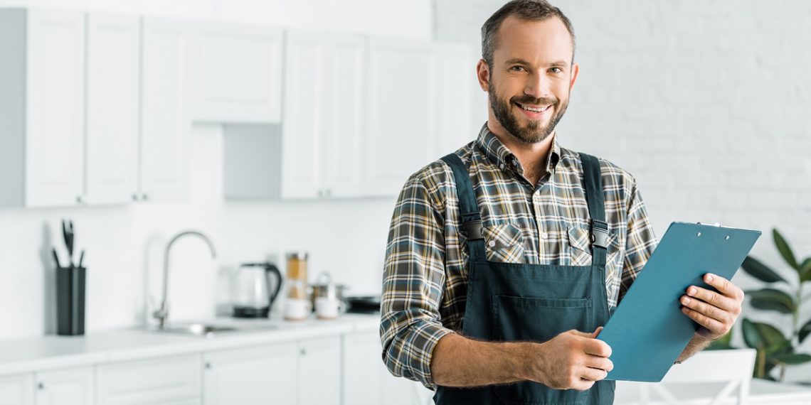 smiling handsome plumber holding clipboard and looking at camera in kitchen