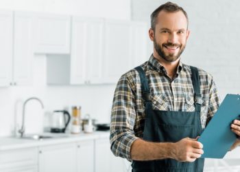 smiling handsome plumber holding clipboard and looking at camera in kitchen