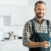 smiling handsome plumber holding clipboard and looking at camera in kitchen