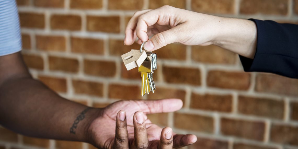 A close-up shot of a real estate agent or property manager dropping a set of house keys into the open palm of a new homeowner or tenant. The keys feature a small wooden house-shaped keychain, set against a blurred red brick wall background.