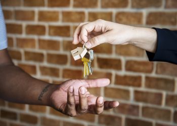 A close-up shot of a real estate agent or property manager dropping a set of house keys into the open palm of a new homeowner or tenant. The keys feature a small wooden house-shaped keychain, set against a blurred red brick wall background.