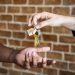 A close-up shot of a real estate agent or property manager dropping a set of house keys into the open palm of a new homeowner or tenant. The keys feature a small wooden house-shaped keychain, set against a blurred red brick wall background.