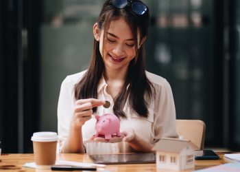 mortgage, investment, real estate and property concept - close up of woman putting money into pink piggy bank for loan or save for buy a new house.