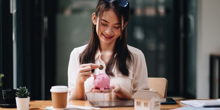 mortgage, investment, real estate and property concept - close up of woman putting money into pink piggy bank for loan or save for buy a new house.