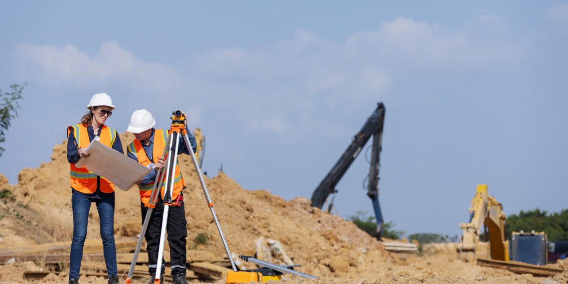 Two construction workers in safety vests and hard hats on a dirt construction site. One person holds a large blueprint while the other operates a surveying level mounted on a tripod. In the background, yellow excavators and a blue sky are visible.