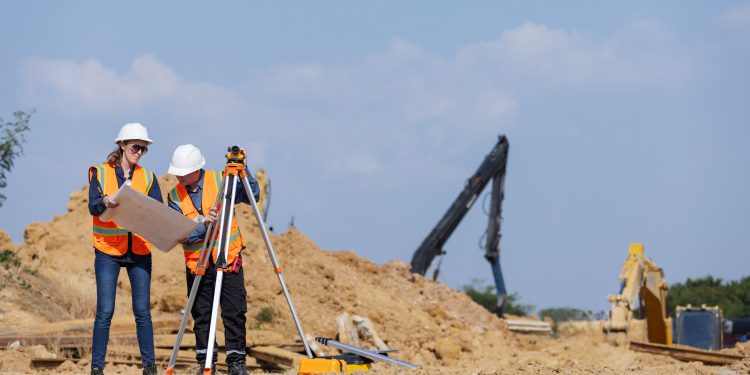 Two construction workers in safety vests and hard hats on a dirt construction site. One person holds a large blueprint while the other operates a surveying level mounted on a tripod. In the background, yellow excavators and a blue sky are visible.
