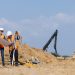 Two construction workers in safety vests and hard hats on a dirt construction site. One person holds a large blueprint while the other operates a surveying level mounted on a tripod. In the background, yellow excavators and a blue sky are visible.