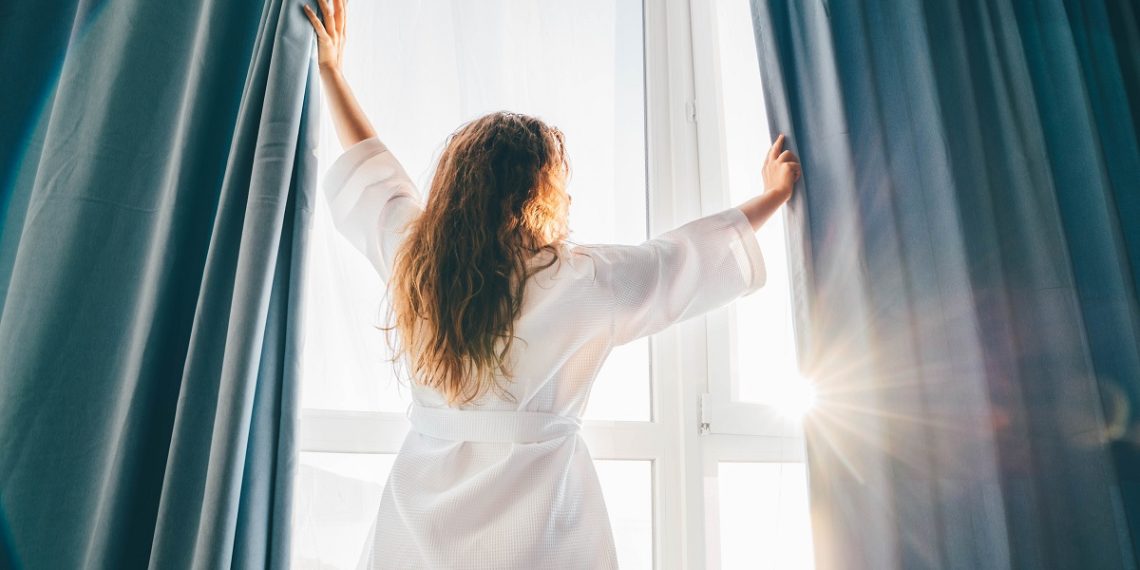 woman-opening-curtains-in-room-at-sunrise
