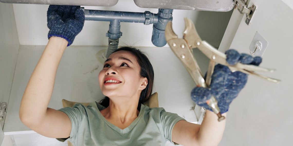 Cheerful young woman wearing protective gloves when fixing leaking sink pipe in kitchen