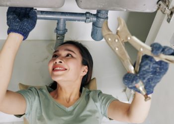 Cheerful young woman wearing protective gloves when fixing leaking sink pipe in kitchen