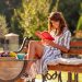 Beautiful young woman enjoying sunny summer morning outdoors, drinking coffee and reading a book on the backyard terrace