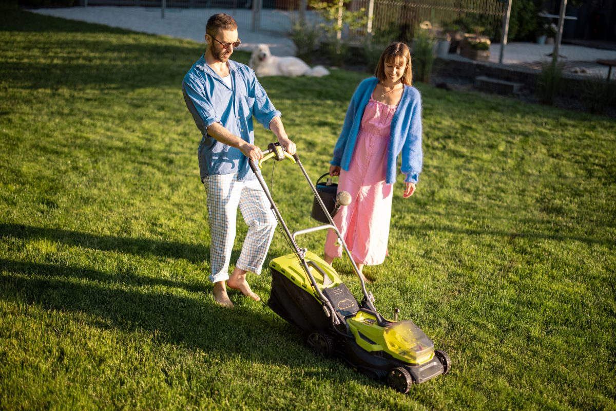 Man mows the lawn with lawn mower, spending summer time with his wife at backyard of country house. Young family take care of garden on summer evening